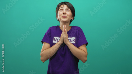 Obraz na plátně Woman with hands pressed together in a pleading prayer gesture wearing a volunteer t shirt against teal backdrop in a studio; hopeful gratitude
