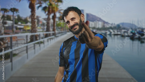 Fotografía Man in soccer jersey shows middle finger while smiling on a street side marina pier with palm trees and boats; defiance