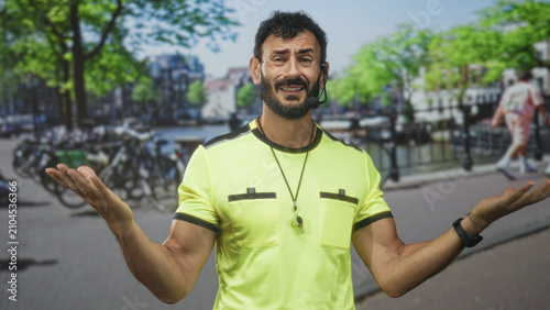 Photos Man referee in neon yellow shirt wearing whistle and headset, open palms shrug gesture on a city street by canal and parked bikes; confusion