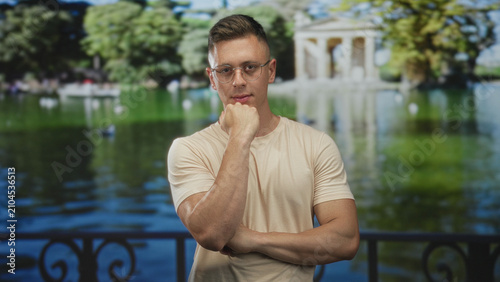 Bild auf Leinwand Man with hand on chin and folded arm posing by lakeside railing in front of a classical temple building, wearing glasses and beige t shirt; contemplation calm