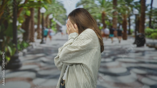 Obraz na plátně Woman with hands covering face on a palm lined street, wearing striped shirt and jeans, side profile visible; anxiety vulnerability