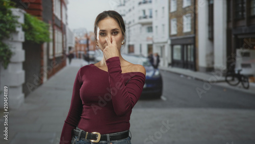 Photos Woman pointing finger at camera on a quiet urban street, wearing maroon long sleeve sweater, jeans and belt, earrings visible; confidence direct address