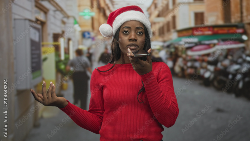 Fototapeta premium Woman in santa hat speaking into smartphone and waving hand on a crowded market street; holiday greeting cheerful.