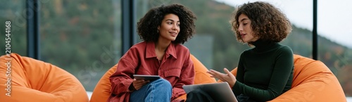 Two young professionals having a casual meeting on orange beanbag chairs in a modern office with mountain view for coworking banner background