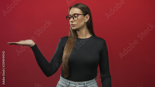 Cuadro en lienzo Woman with glasses holding palm up and raising index finger in a red studio pointing pose, denim waist visible; assertive instruction