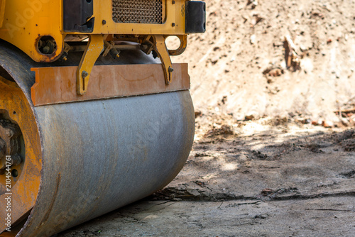 Close-up view of the front, smooth, metal drum of a heavy yellow construction machine, a road roller, parked on uneven, sandy-gravel ground at a construction site under harsh, natural sunlight.