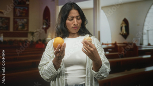 Foto Woman holding an orange and a muffin in her hands, frowning while comparing the foods inside a church; hesitation choice restraint
