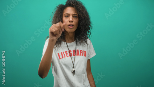 Obraz na plátně Woman lifeguard pointing finger with raised hand and whistle on lanyard in studio; concern vigilance