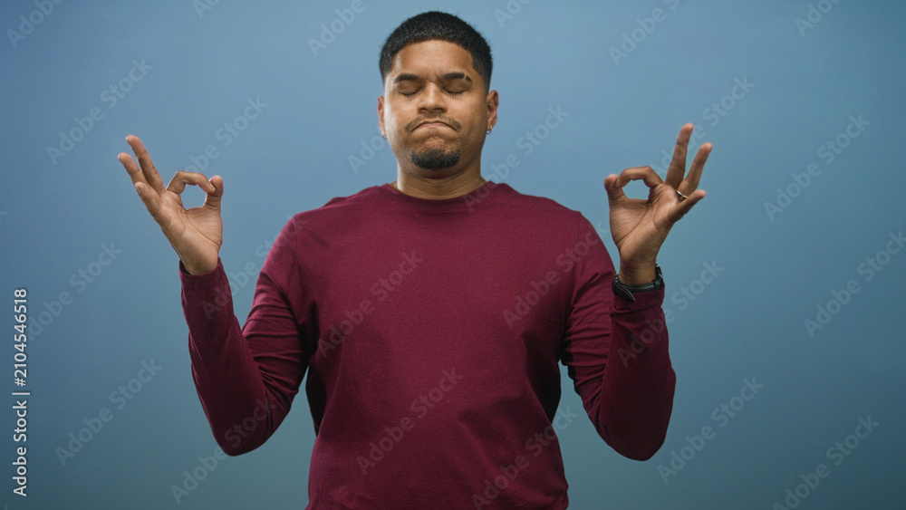 Fototapeta premium Man with hands pressed together praying in studio, eyes closed and wearing maroon sweater; calm reflection.