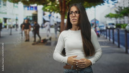 Принти на полотні Woman covers eyes with hand on busy street wearing white shirt and jeans with visible belt and long hair; embarrassment avoidance
