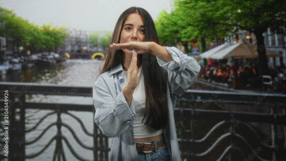 Fototapeta premium Woman makes time-out t gesture with hands on street canal bridge by riverside railing and outdoor cafe; confidence.