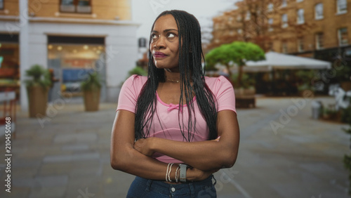 Canvastavla Woman in pink top with crossed arms showing forearms and a smartwatch, standing in a street plaza with cafe umbrellas and planters; annoyance defiance