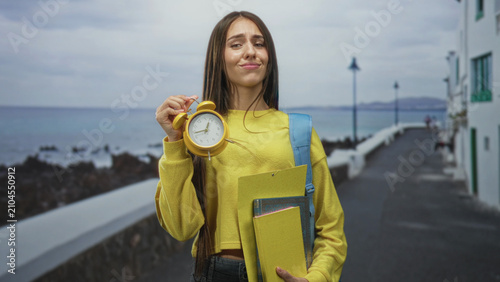 Принти на полотні Teenage student holds a yellow alarm clock and school folders on a street by the sea, lifting the clock with a smirk and raised wrist gesture; time pressure playful