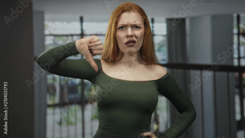 Φωτογραφία Redhead woman with bare shoulder giving thumbs down gesture in a modern building lobby wearing green off shoulder top; disapproval critique
