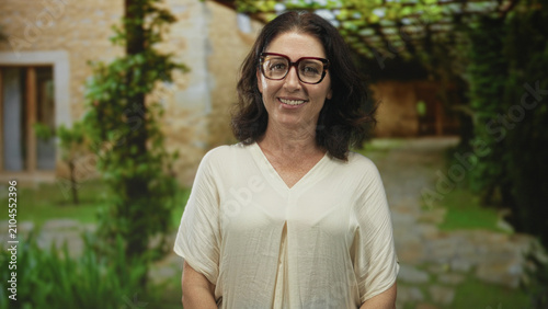 Foto Woman wearing glasses smiling at camera in building courtyard with white blouse visible and relaxed pose framed by pergola; contentment reflection