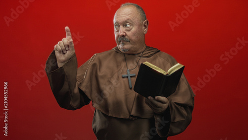 Foto Man senior monk holds bible in left hand and points right index finger, wearing brown habit and cross necklace in studio with red backdrop; solemn authority