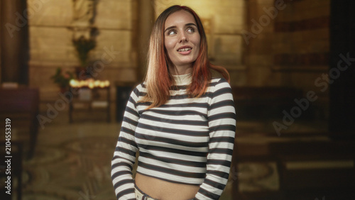Bild auf Leinwand Young redhead woman smiles and tilts head, wearing a striped crop top and belt, standing inside a church building; contentment