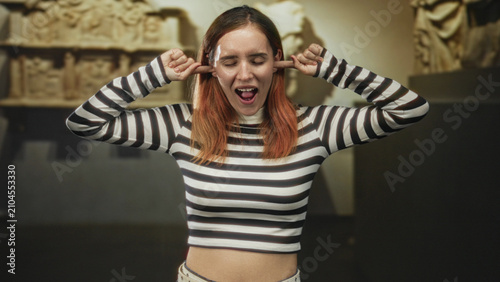 Obraz na plátně Young woman plugs ears with fingers in museum, wearing a striped crop top and grimacing with eyes closed, overwhelmed by loud noise and bright lighting; frustration