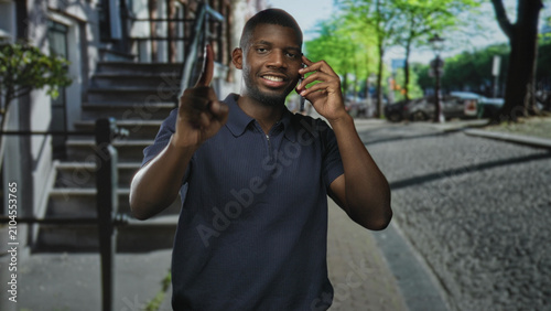 Photos Young black man on street holding smartphone to ear and pointing index finger, smiling near stoop and parked car; confidence communication