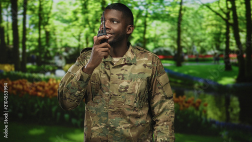 Foto Man soldier speaks into radio in a forest wearing camouflage uniform, holding a handheld transceiver and scanning the area with a slight smile; duty vigilance