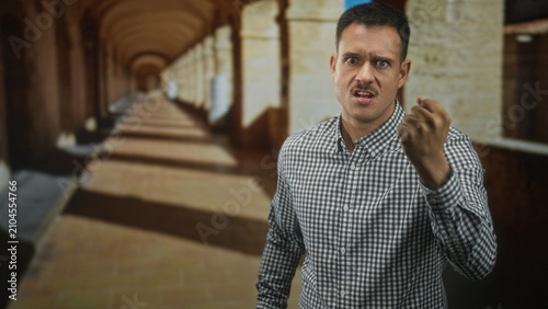 Quadro su tela Young hispanic man with moustache clenches fist forward in arched building corridor wearing check shirt; anger defiance tension