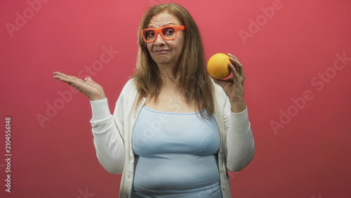 Fotografie Woman holding an orange in right hand and shrugging with left palm up, wearing orange glasses and light blue tanktop against a pink studio backdrop; wry bemusement
