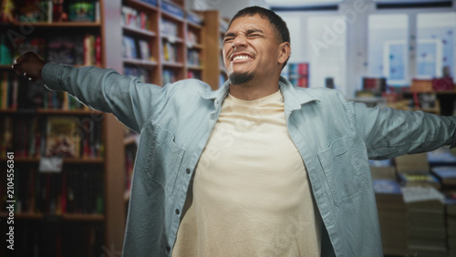 Fotografie Man stretching arms wide while holding dollars in hand inside a bookstore, smiling with closed eyes and casual jacket; celebration success
