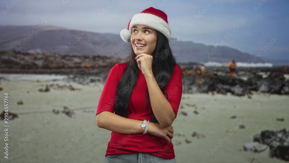 Obraz premium Young woman wearing santa hat smiles with hand on chin and crossed arm in a studio beach set; holiday joy.