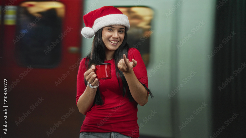 Obraz premium Woman in a santa hat holding a red mug and beckoning with her hand on a subway platform inside a building; holiday joy.