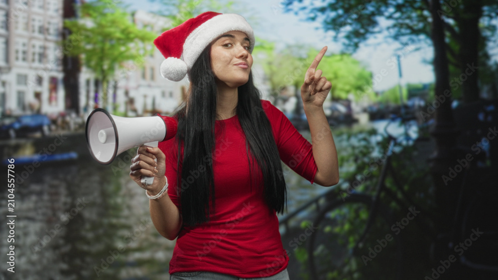 Obraz premium Young woman wearing santa hat holds a megaphone and points finger on a city street by canal buildings, smiling and addressing unseen crowd; holiday cheer.