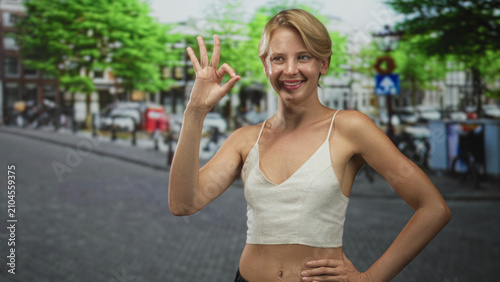 Tableau sur toile Caucasian blond woman shows ok with hand and puckered lips on street in urban cobblestone scene wearing cami and casual top; playful confidence