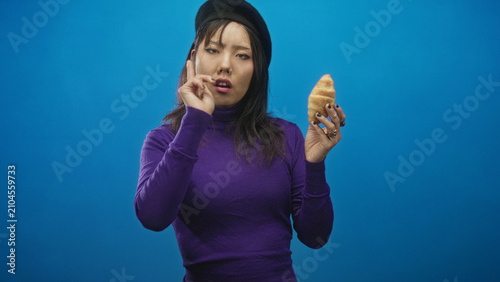 Obraz na plátně Woman in purple turtleneck and beret holding croissant and pointing finger with hand forward in studio; quizzical confusion