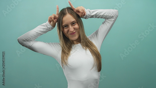 Fotografi Young woman smiling and making horns gesture with fingers to head in studio wearing white long sleeve top against teal backdrop; playful mischief