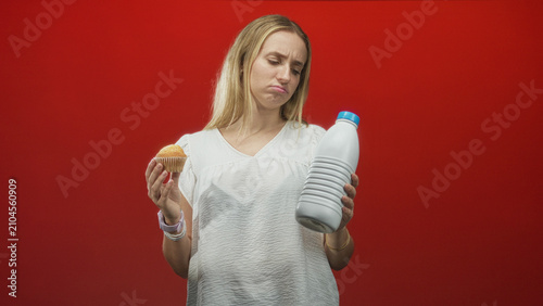 Canvas Print Woman holding muffin and large milk bottle, pouting lips, bare hands and white blouse against red studio backdrop; indecision