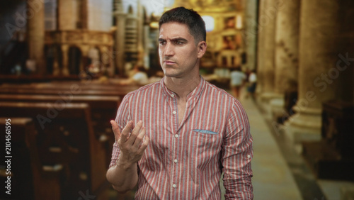 Fotografie Young hispanic man hand on chin in church nave with striped shirt and wooden pews visible, pensive expression; contemplation thoughtful
