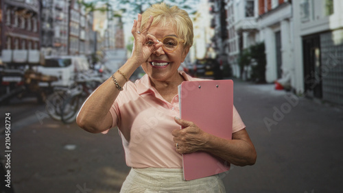 Bilde på lerret Woman holding pink clipboard grips folder and makes ok sign to eye with her hand on a street near bicycles and shops; joyful confidence
