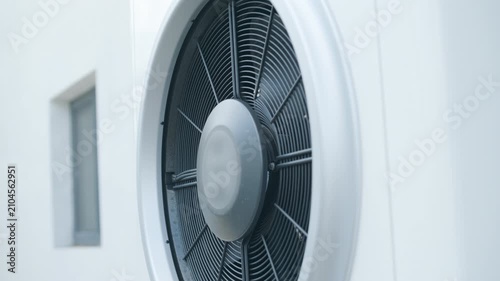 Modern outdoor air-source heat pump unit with a large fan on a white residential building wall, low angle view.
