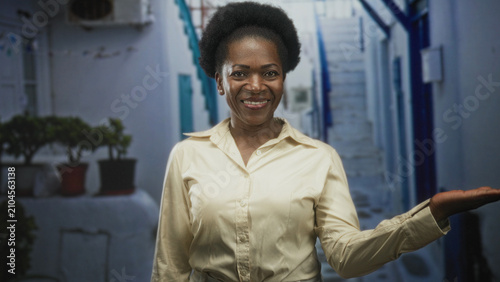 Φωτογραφία Woman smiling and presenting an open right hand palm while wearing a beige shirt in a narrow white street with stairs and potted plants; friendly