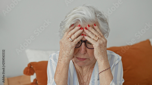 Photos Elderly woman sitting on bed holding her head with hands, glasses askew and red nails visible in building bedroom scene; distress headache