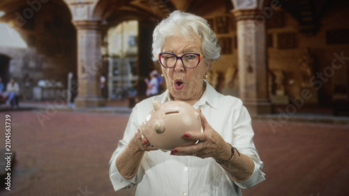 Fotografija Senior woman wearing glasses carefully receives a ceramic piggy bank with both hands in a historic building arcade; surprise savings
