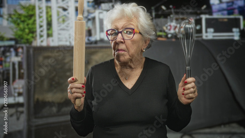 Tableau sur toile Elderly woman wearing glasses and black sweater holds rolling pin and whisk with raised hands in studio set, red nails visible; determination