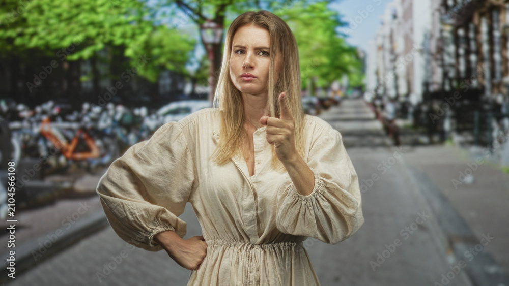 Fototapeta premium Blonde woman in linen dress standing with hands on hips and one finger raised on a street with bicycles and buildings behind her; determination.