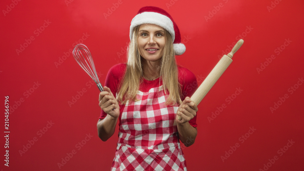 Fototapeta premium Woman holding whisk and rolling pin, smiling with mouth open, wearing santa hat and checked apron in studio; holiday baking joyful.