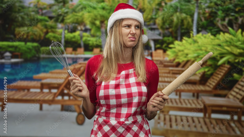 Fototapeta premium Woman wearing santa hat and gingham apron holds whisk and rolling pin, grimaces while standing poolside by a hotel building; holiday frustration.