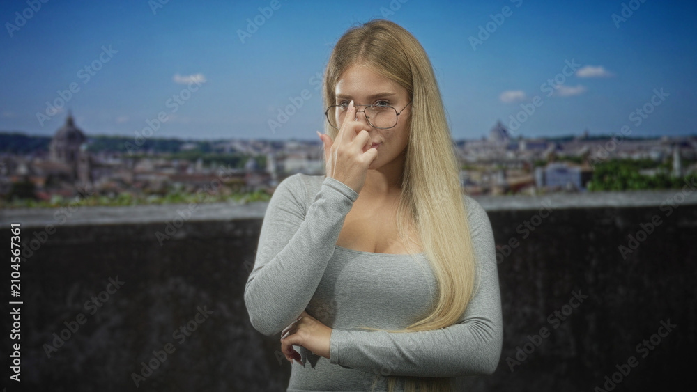 Obraz premium Woman touching glasses with finger at nose bridge on building terrace overlooking city; thoughtful contemplation.