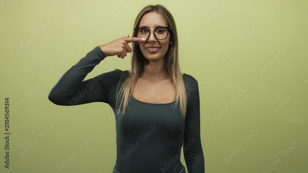 Fototapeta premium Woman points finger to nose while smiling and wearing glasses in studio with olive green wall; playful.