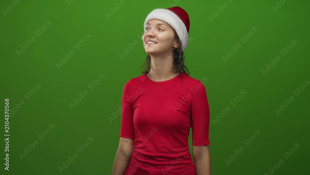 Fototapeta premium Woman in santa hat and red shirt smiles and tilts her head to the right in studio green screen setting; cheerful holiday spirit.