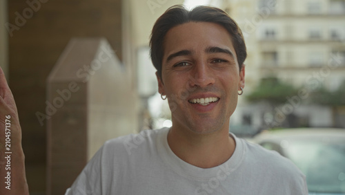 Tableau sur toile Man smiling with face forward, left hand on doorway, wearing white tshirt and hoop earring on street near building entrance; casual warmth