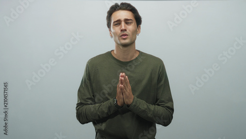 Картината върху платно Man with hands pressed together in prayer gesture, closed eyes and pleading lips, wearing olive long sleeve shirt and wristwatch against a neutral grey studio backdrop; plea hope