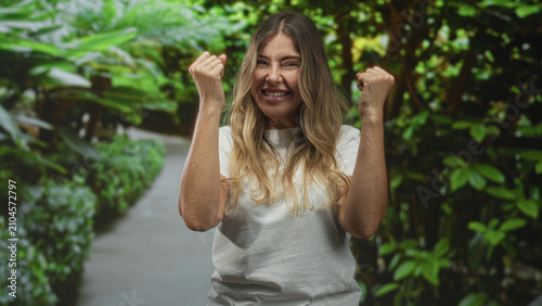 Foto Middle age hispanic woman with clenched fists smiling on a forest path in white tshirt and long hair visible, celebrating a small victory; joy triumph